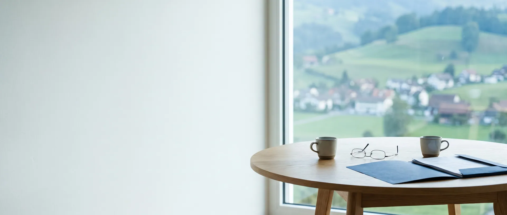 Runder Holztisch mit zwei Tassen Tee und Ordner vor einem Fenster mit Hügelblick
